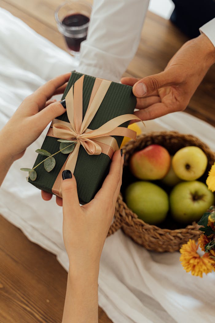 A close-up of hands exchanging a green gift next to a fruit basket on a wooden table.